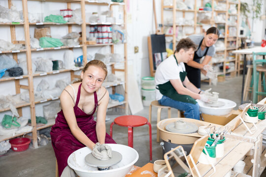 Young woman teacher in apron helps teenager boy and teenager girl students to make product from clay on potter's wheel