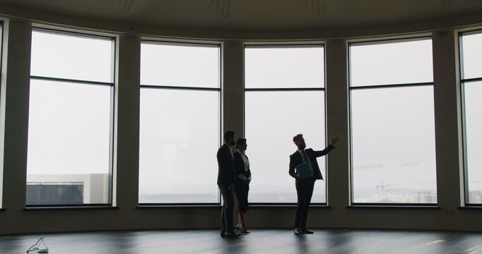 Expert realtor presenting to business professionals spacious office with large panoramic windows. Business people exploring options for purchase or rent in modern space with cityscape in background.