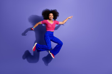 Young cheerful afro haired woman in vibrant outfit jumps elegantly against a solid purple background