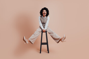 Confident businesswoman in stylish attire sits on a stool with a joyful expression against a beige...