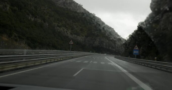 A subjective shot taken from inside the vehicle shows the motorway between the mountains in the rain. The windscreen wipers can be seen passing through the glass.