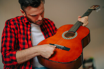 Man in a red plaid shirt tuning an acoustic guitar, focused on adjusting the strings. Indoor...