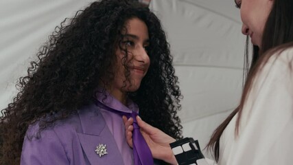 Handheld shot of female stylist chatting while adjusting tie on friendly young biracial female model wearing lilac designer suit preparing for commercial photo shoot in professional studio