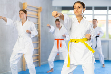 Concentrated teenage girl in white kimono practicing punches and blocks in gym during martial arts workout with female group. Shadow fight, combat sports training concept ..