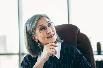 Confident mature businesswoman with gray hair in office chair wearing formal attire and glasses,...
