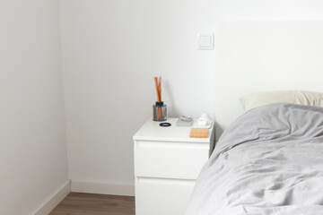 Minimalist aesthetic bedroom interior. White bedside table with aroma diffuser, charging cable, cosmetic container, phone stand, wireless charger and hair tie