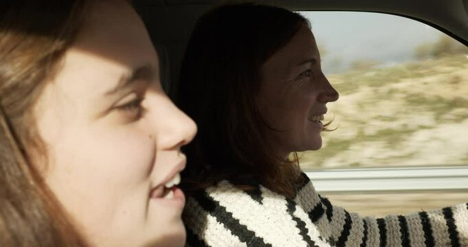 A close-up of mother and daughter in a car. The mother is driving. They look at each other with complicity and smile. The sun is shining. The background is a field with grey clouds. Focus on the mothe