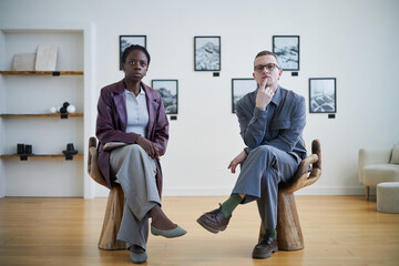 African American woman and Caucasian man sitting on wooden stools in modern art gallery, with framed photos on white walls, focusing on art, bright room, minimalist design