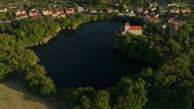 Full aerial view of the Wehrkirche Beucha perched on a cliff above a former quarry lake, surrounded by forest and village houses. Captured in warm evening light near Leipzig, Germany.