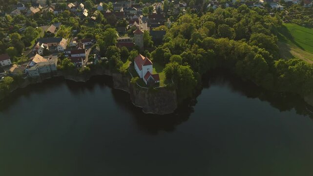 Ascending and backward drone shot of the Wehrkirche Beucha in Saxony, Germany. The white church stands atop a steep rock above a quarry lake, captured in soft golden evening light.