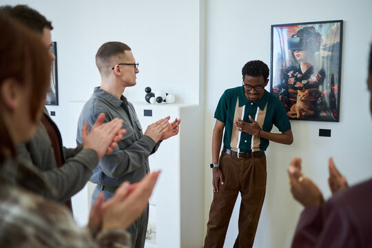 Group of people applauding a man standing in modern art gallery. Man showing gratitude while surrounded by diverse individuals appreciating contemporary art installation