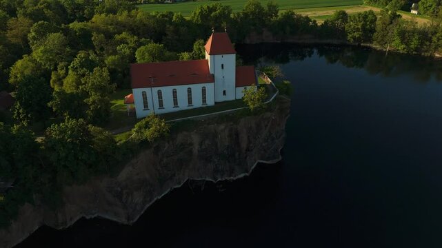 Smooth lateral drone flyby of the Wehrkirche Beucha, perched on a rocky cliff above a quarry lake in Saxony. Captured in soft sunset light, showcasing the dramatic edge and surrounding forest.
