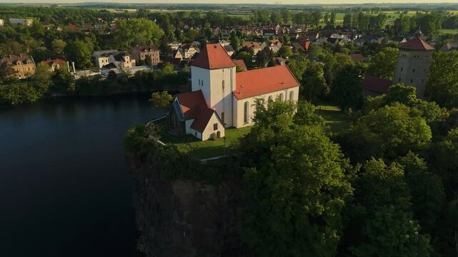 Golden hour drone shot of the Wehrkirche Beucha, a medieval church perched on a sandstone cliff above a lake. The light emphasizes the red roof and white walls, surrounded by forest and village.