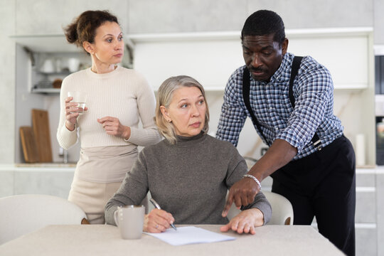 Thoughtful old woman compiling testament while middle-aged man and woman competing with each other in the kitchen
