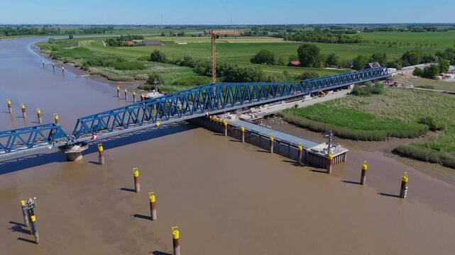 Aerial view of the new Friesenbr&uuml;cke railway bridge over the Ems River in Germany. The swing-lift mechanism allows ship traffic to pass. Construction scene with piers, crane, and rural surroundings.