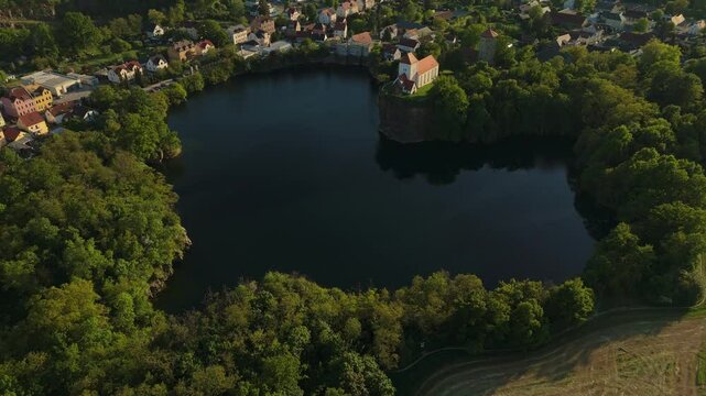 Drone clip of the Wehrkirche Beucha on a cliff above a heart-shaped quarry lake in Saxony, Germany. Surrounded by forest and village houses, captured in golden hour light.