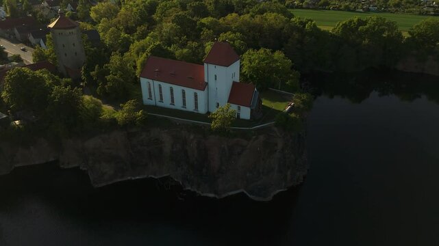 Cinematic drone shot of the Wehrkirche Beucha near Leipzig at sunset. The historic church sits dramatically on a cliff above the flooded quarry, surrounded by forest and glowing evening light.