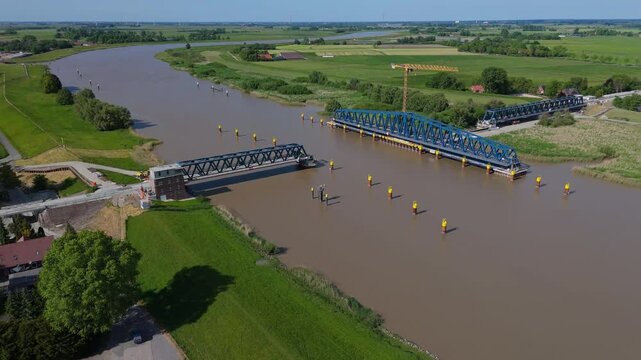 Wide drone shot of the Friesenbr&uuml;cke in open position during construction over the Ems River in Germany. Highlights the rotating section, ship passage, and rural surroundings on a clear summer day.