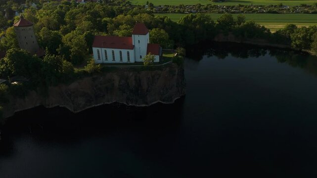 Aerial orbit of the historic Wehrkirche Beucha perched atop a steep cliff at sunset, surrounded by forest and lake. Captured near Leipzig, Germany.