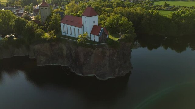 Stunning aerial view of the Wehrkirche Beucha, perched on a high cliff above a flooded quarry. The medieval church glows in the warm sunset light, surrounded by forest and tranquil waters.