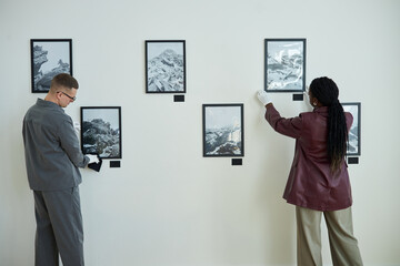 Two people arranging monochrome photographs on wall of an art exhibit, using gloves to handle...