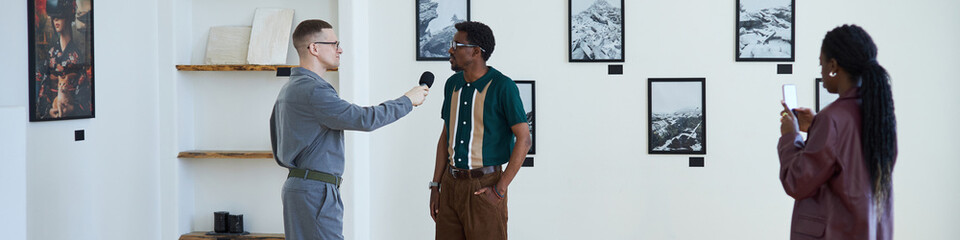 Header with black man standing in an art gallery, being interviewed by a man holding microphone...