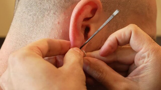Close-up of a professional piercer piercing a man's ear with a sterile needle. The client's ear is held gently for precision. The scene demonstrates hygienic conditions and meticulous technique. 4k