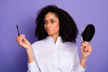 Young woman in white shirt holding mascara wand and mirror against purple background expressing...