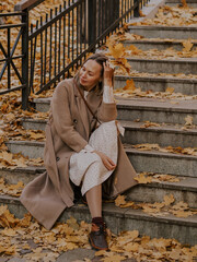 Beautiful blonde young woman in hat and coat sitting on the stairs in the autumn park.