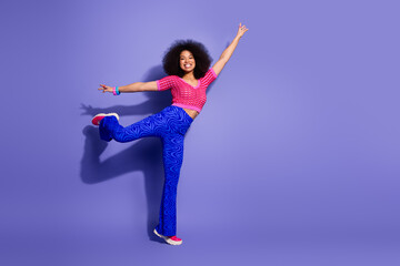 Joyful young woman in vibrant retro outfit posing against a purple background, expressing energy...