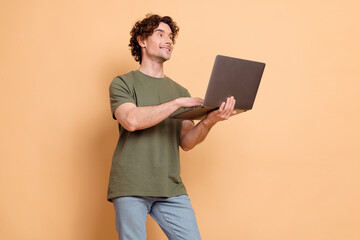 Young stylish man with curly hair holding a laptop against a beige background showcasing a casual...