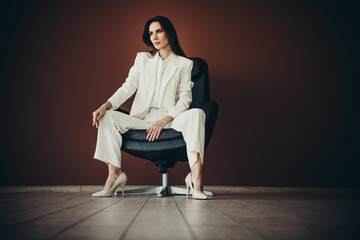 Beautiful businesswoman in white formalwear sitting on a chair against a stylish brown background...