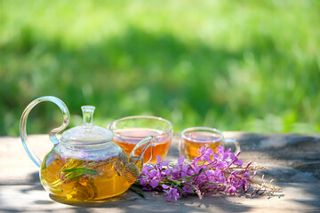 Glass teapot and cups with tea from flowers of fireweed on table in garden, abstract natural...
