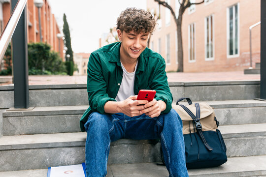 Young student with curly hair using red mobile phone app in University campus