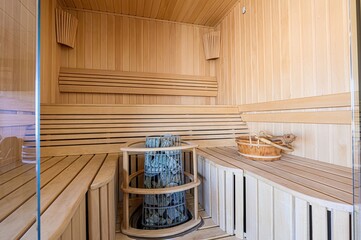cozy sauna featuring light wood paneling, tiered benches, and a stone heater. A wooden bucket and accessories add to the relaxing atmosphere