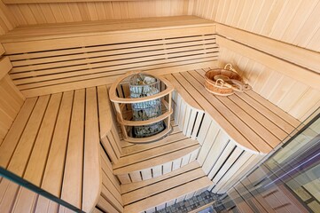 cozy sauna featuring light wood paneling, tiered benches, and a stone heater. A wooden bucket and...