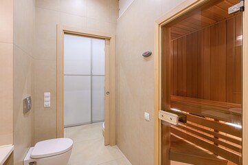 modern bathroom with beige tile walls, featuring a white toilet and two doorways. One leads to a shower with sliding doors, the other to a sauna with a glass door