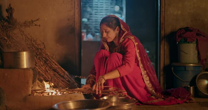 Indian villager gen x woman preparing food using traditional earthen mud stove indoor house Asian young female wear red saree cooking on open fire flam inside kitchen home