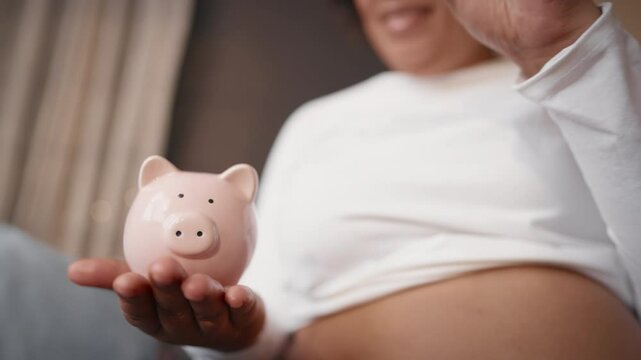 Pregnant woman saving money for future baby, closeup of piggy bank and belly. African american lady putting coins, careful mother waiting for child, big abdomen in eight month pregnancy, finance