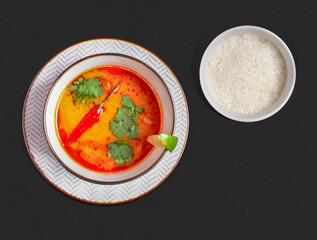 Top-view photograph: traditional Thai tom yum soup with vibrant red-orange broth, garnished with...