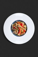 Top-view photograph: Asian stir-fried udon noodles with vegetables, served in a deep plate with a blue textured center and wide white rim, on a dark background. 