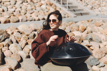 young caucasian woman playing black handpan with hands, ethnic metal drum instrument, sits on stones of enbankment, beautiful relaxing music for meditation