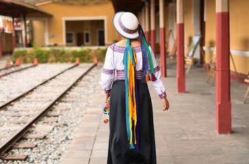 Indigenous woman wearing traditional andean clothing and hat walks along a train station platform,...