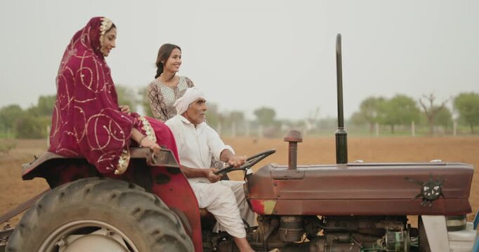 Indian village farmer young woman lady wear red saree smiling sitting on tractor outdoor Asian mother with teen age girl talk gossip old aged male man driving outside farm rural area enjoy desi life	