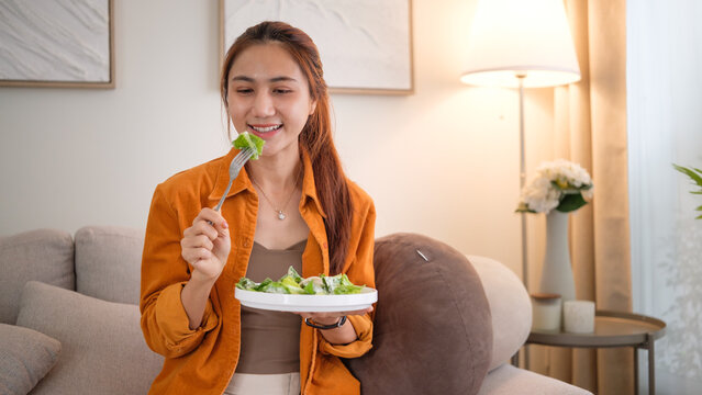 Relaxed lifestyle moment — woman eating a fresh vegetable salad in a warmly lit modern living room. - Powered by Adobe