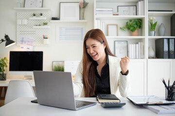 Smiling woman raising her fist in triumph while looking at her laptop, symbolizing achievement, good news, or professional victory.