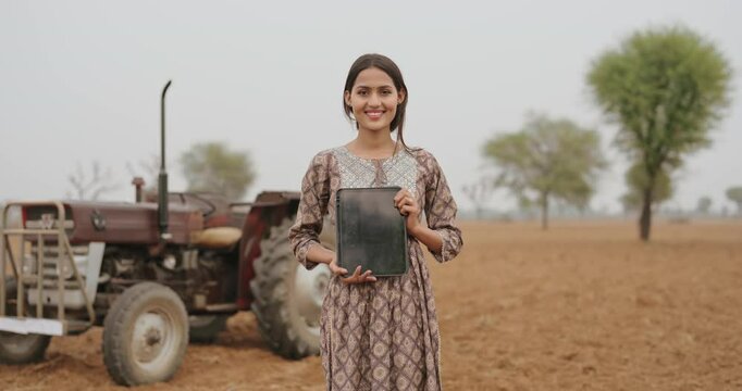 Indian young adult gen z farmer woman lady wear casual dress standing outdoor farmland holding tablet device looking at camera asian smiling teen age girl do pose staring cam outside desi life