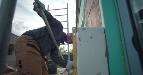 Builder working on insulation installation, kneeling on scaffolding, using level tool for precision, focused on exterior wall of residential house under construction