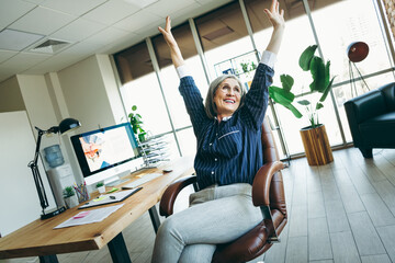 Confident businesswoman with grey hair celebrating success in a modern bright office interior with...