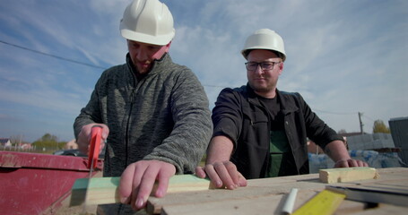 Two builders working together, one using a handsaw to cut wood while other watches, teamwork on...